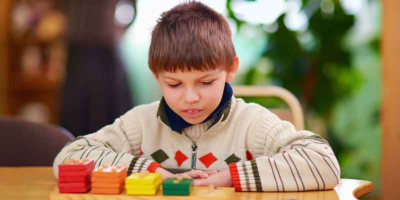A young boy looks down at stacks of wooden squares, which are arranged by color and size across a pegboard.